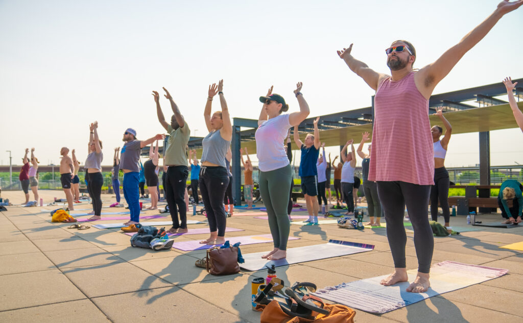 Yoga class participants stand with outstretched arms on a plaza